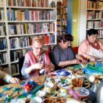 Na zdjęciu seniorki siedzą przy stole i wykonują wiozenne wianki. Na stole przed nimi kolorowe wstążki, nożyczki, niebieski koszyczek paluszkiw filiżance w żółto zielone wzory, dwa srebrne termosy oraz ciasteczka drobne na szklanej paterze.