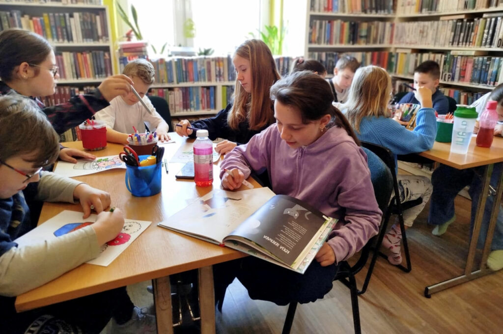 na zdjęciu widać dzieci siedzące przy stołach w sali bibliotecznej i wykonujące prace plastyczne
