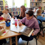 na zdjęciu widać dzieci siedzące przy stołach w sali bibliotecznej i wykonujące prace plastyczne