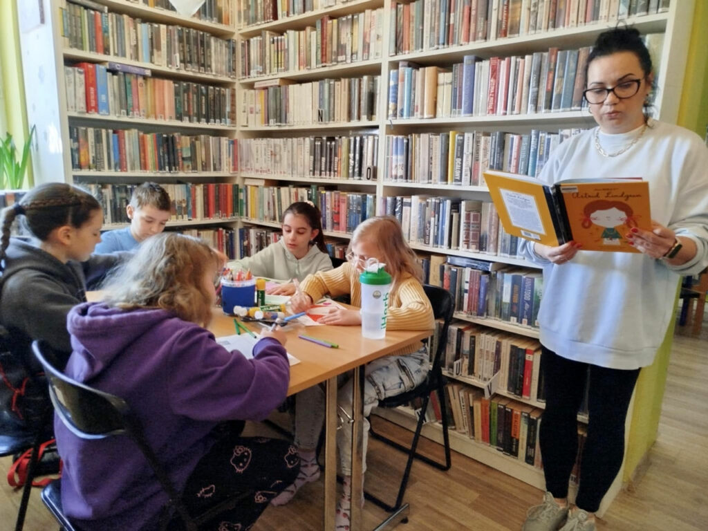 Na zdjęciu Pani bibliotekarka stoi przy regale z książkami trzyma w reku książkę i czyta ją. Z lewej strony przy stoliku siedzi piątka dzieci i wykonuje pracę plastyczną. Na stole kartki, kredki kleje