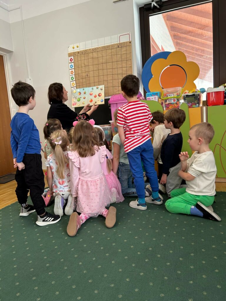 Na zdjęciu bibliotekarka siedzi na krześle i pokazuje dzieciom książeczkę. Wokół niej dwóch chłopców stoi, ósemka przedszkolaków siedzi na podłodze zwrócona do pani bibliotekarki, która pokazuje ilustracje w książce.