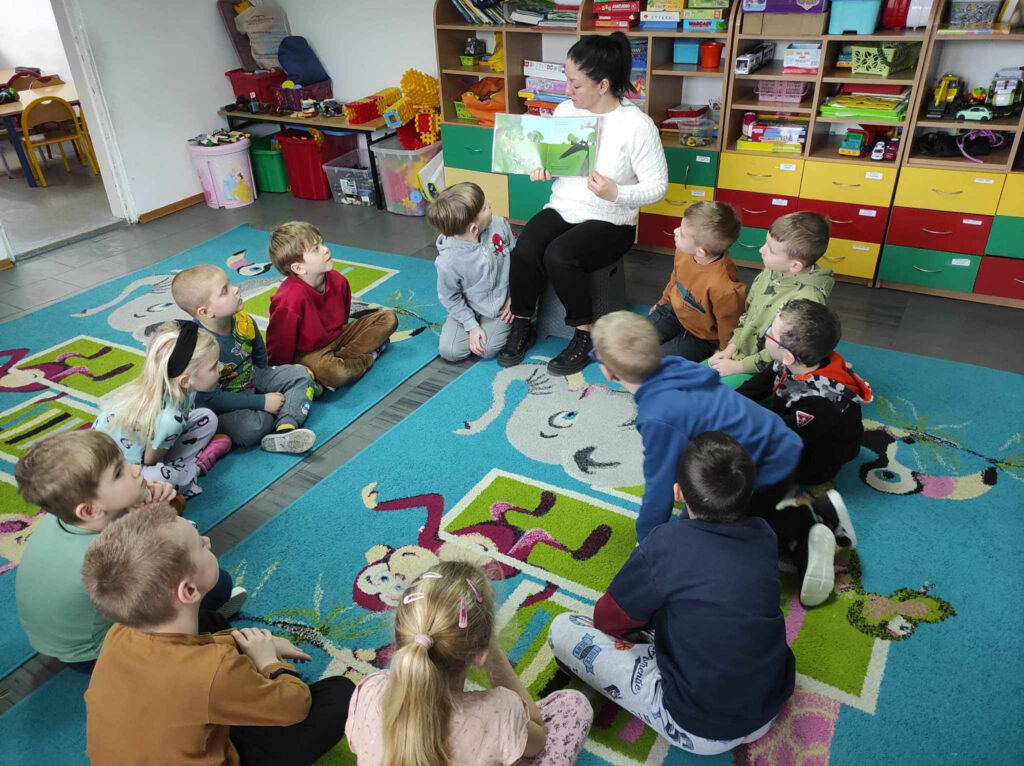 na zdjęciu widać salę przedszkolną, w kole na dywanie siedzą dzieci, bibliotekarz czyta książkę