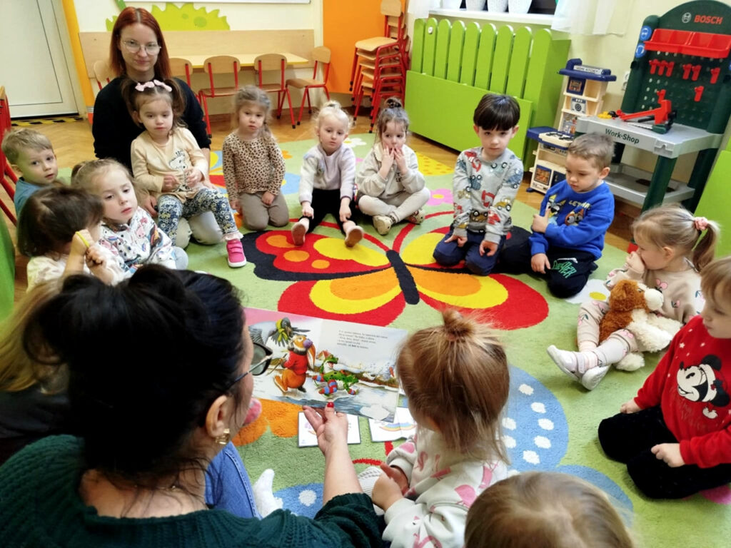 na zdjęciu widać dzieci siedzące w kułeczku na dywanie. Pani bibliotekarka czyta bajeczkę.