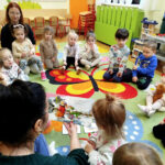 na zdjęciu widać dzieci siedzące w kułeczku na dywanie. Pani bibliotekarka czyta bajeczkę.
