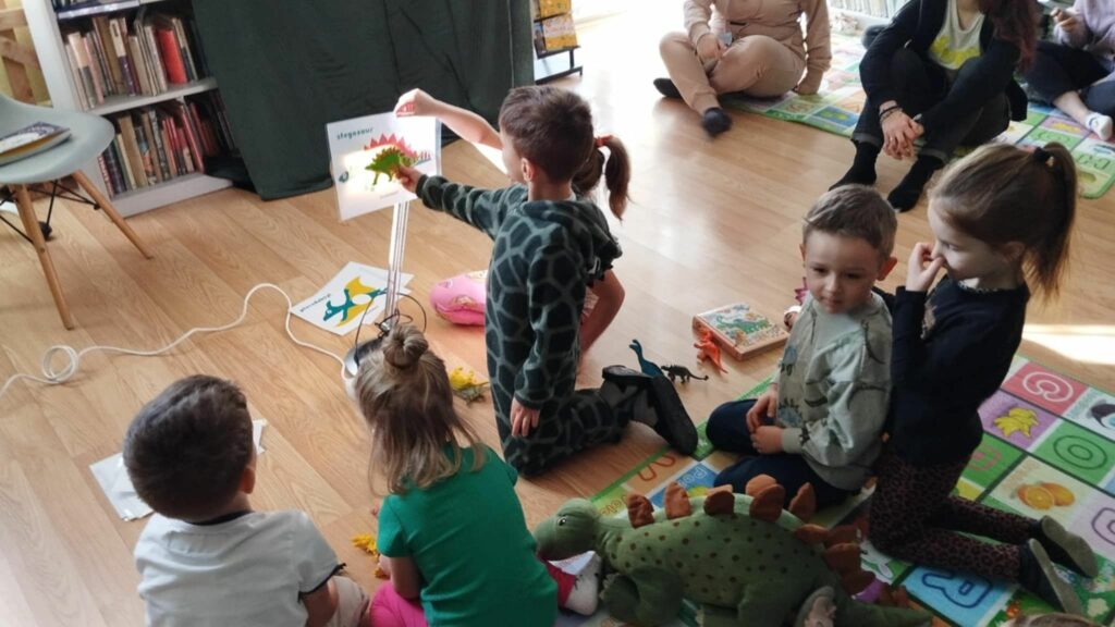 Na zdjęciu dzieci siedzą w bibliotece na podłodze na kolorowych matach