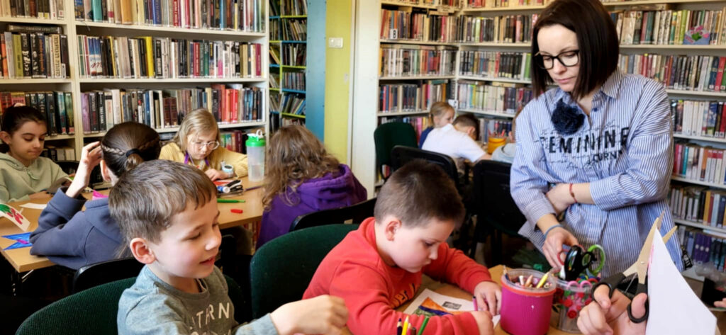 Na zdjęciu widaaaać salę biblioteczną. Przy trzech stołach siedza dzieci i wykonuja prace plastyczne. Przy jednym ze stołów stoi pani bibliotekarka i przygląda się jak dzieci radza sobie z wykonywaniem prac.