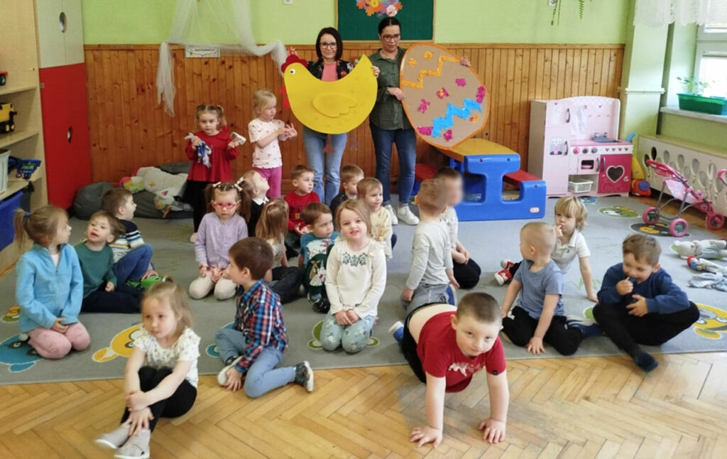 na zdjęciu widać dzieci siedzące na podłodzeza nimi panie trzymaja kurę z filcu