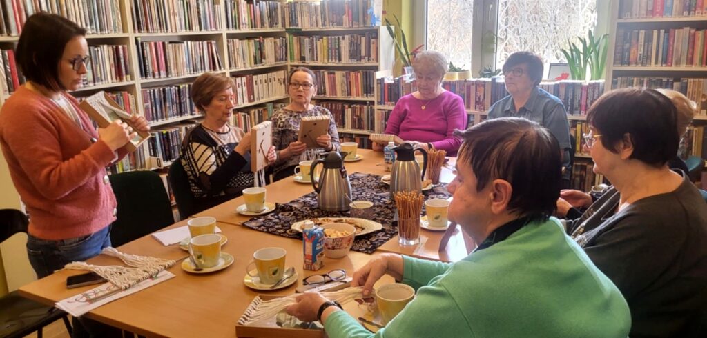 na zdjęciu widać kobiety siedzące wokół stołu i słuchające bibliotekarki która tłumaczy jak wykonać pracę