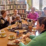 na zdjęciu widać kobiety siedzące wokół stołu i słuchające bibliotekarki która tłumaczy jak wykonać pracę