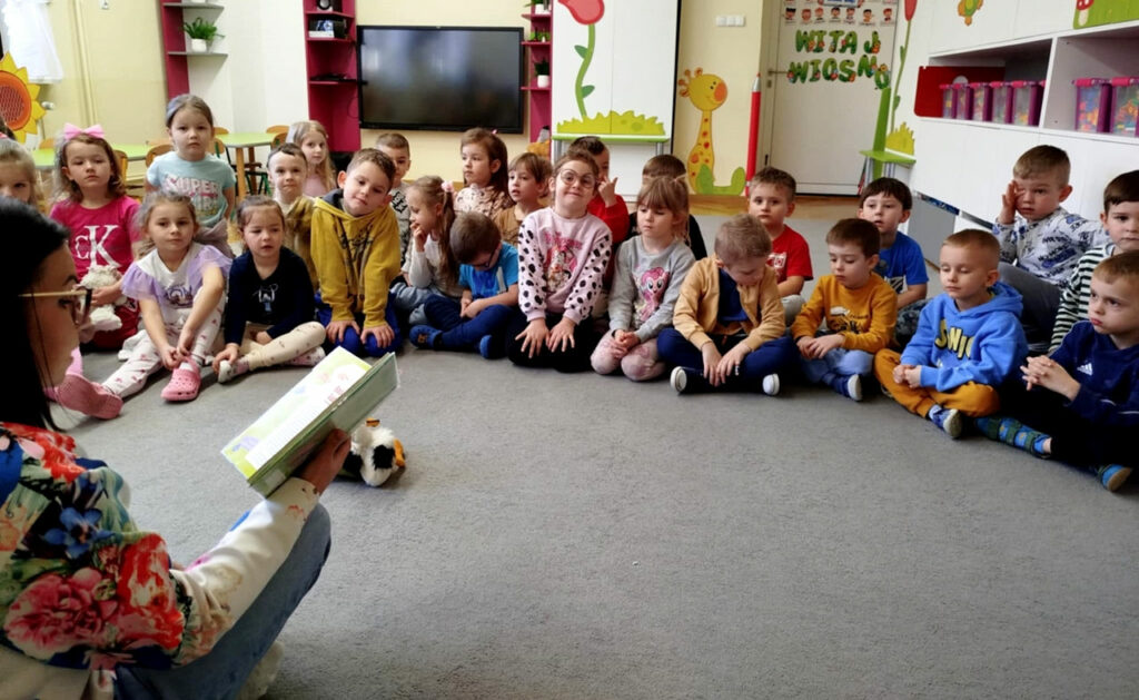 na zdjęciu widać dzieci siedzące na dywanie i słuchające czytanej im książek
