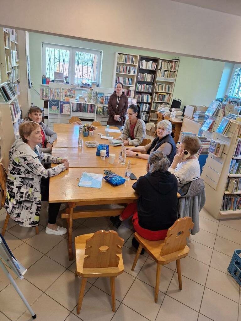 Na zdjęciu kobiety siedzą przy stłach w bibliotece. Wokół regały z książkami.