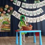 Na zdjęciu widać dziewczynkę w wieku około 10 lat trzyma w ręku mikrofon w tyle napis Wiosenny Konkurs Recytatorski.