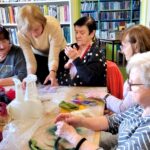 na zdjęciu widać kobiety wokół stołu w sali bibliotecznej a przed nimi kolorowa wełna