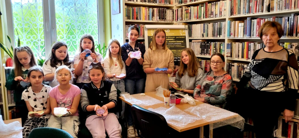 na widać osoby w różnym wieku dzieci i seniorki stojące w sali bibliotecznej