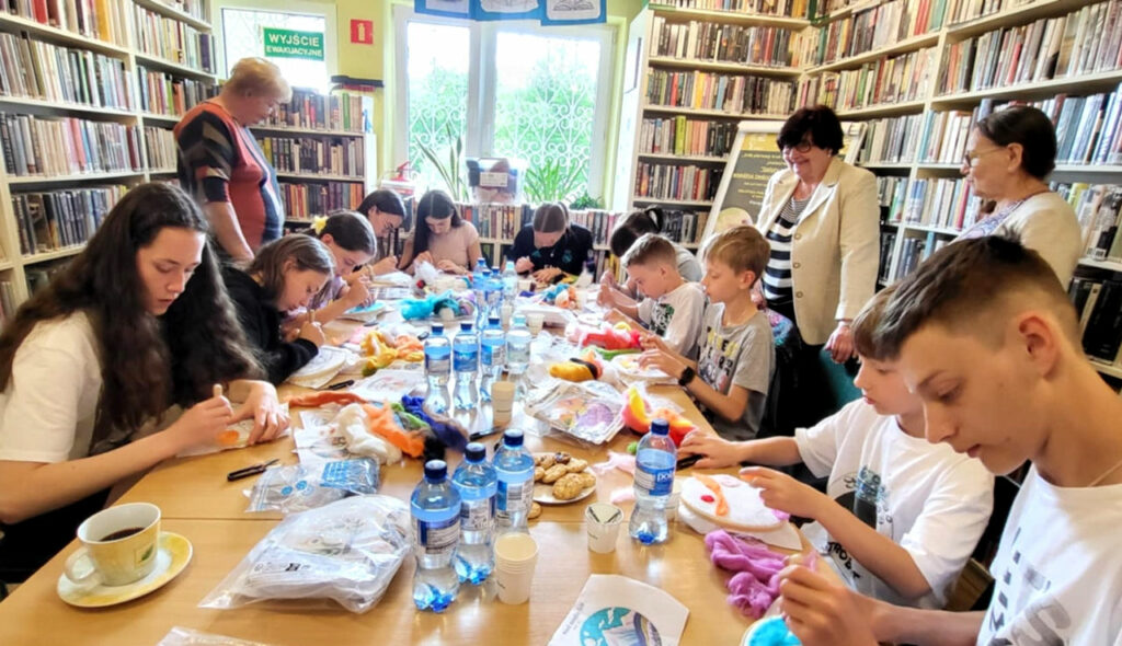 na zdjęciu widać osoby siedzące wokół stołu w sali bibliotecznej i wykonujące obrazy z filcu