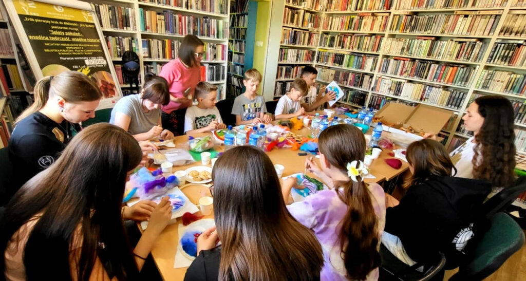 na zdjęciu widać osoby siedzące wokół stołu w sali bibliotecznej i wykonujące obrazy z filcu. Na stole przed nimi materiały plastyczne