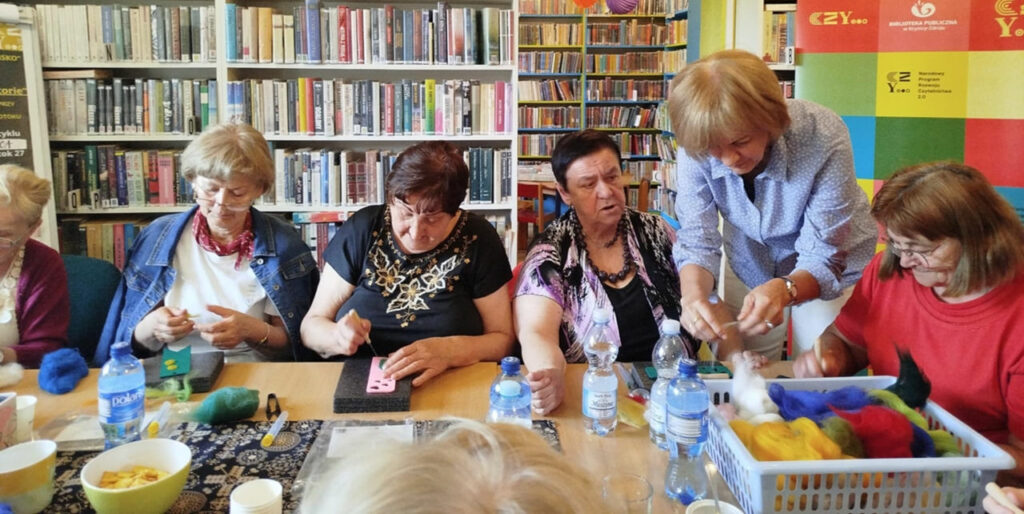 na zdjęciu widać seniorki siedzące wokół stołu w sali bibliotecznej