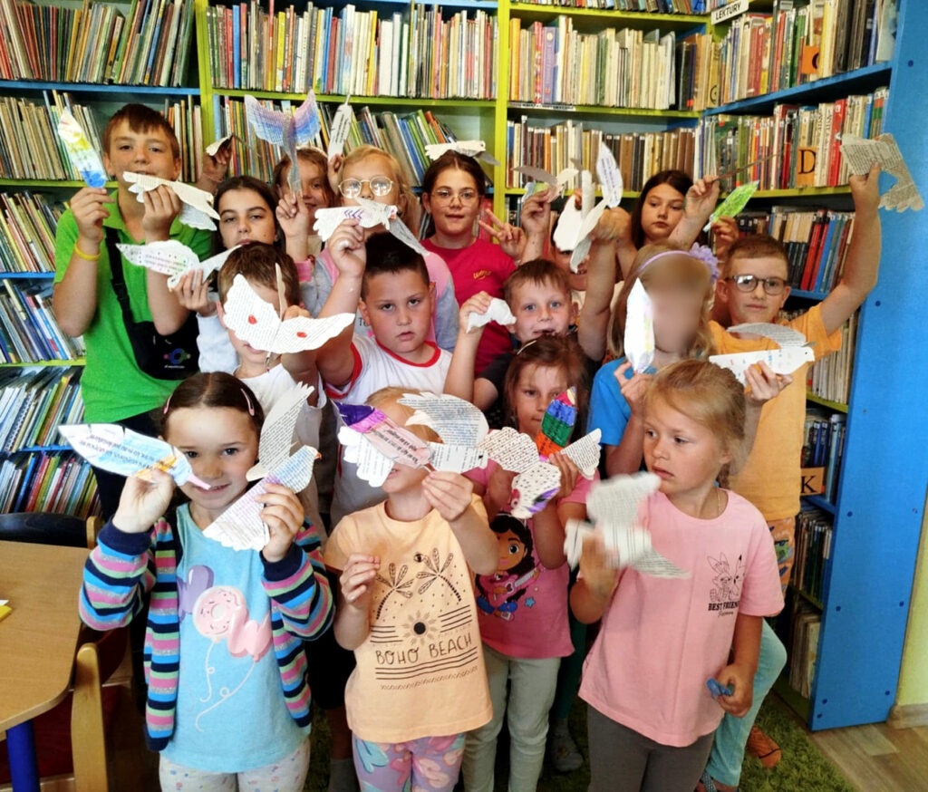 na zdjęciu widać dzieci stojące w sali bibliotecznej. Dzieci trzymają papierowe ptaki