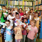 na zdjęciu widać dzieci stojące w sali bibliotecznej. Dzieci trzymają papierowe ptaki