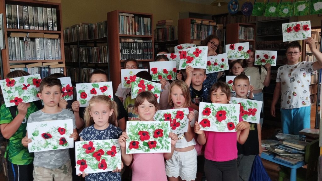 Na zdjęciu dzieci uczestniczące w warsztatach wakacyjnych prezentują ilustracje z makami. Za dziećmi w tyle regały z książkami.