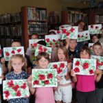 Na zdjęciu dzieci uczestniczące w warsztatach wakacyjnych prezentują ilustracje z makami. Za dziećmi w tyle regały z książkami.