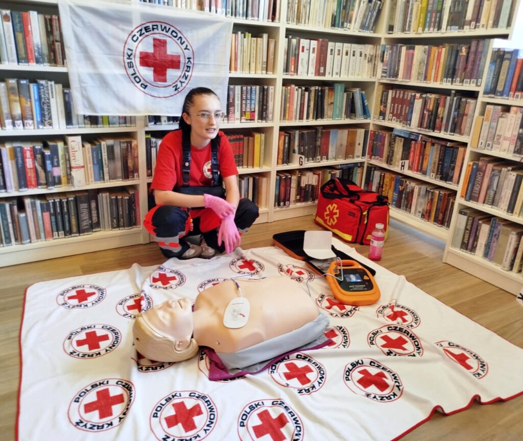 na zdjęciu widać młodą dziewczynę w stroju ratownika. Z tyłu za na regałach z książkami wisi flaga biała a na niej Polski Czerwony Krzyż