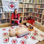 na zdjęciu widać młodą dziewczynę w stroju ratownika. Z tyłu za na regałach z książkami wisi flaga biała a na niej Polski Czerwony Krzyż