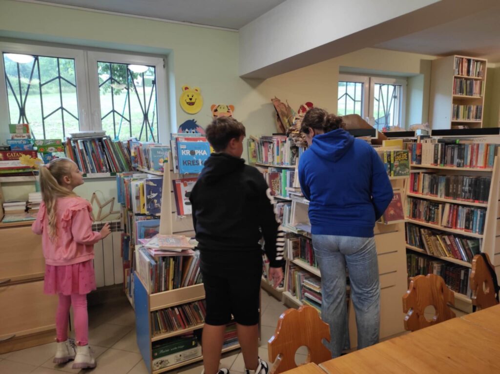 na zdjęciu dwie dziewczynki i chłoiec w sali bibliotecznej oglądaja ksiażki na półkach.