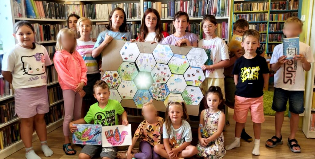 na zdjęciu widać dzieci stojące w sali bibliotecznej. Trzymają wykonane prace plastyczne.