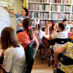 Przy stołach siedza uczestnicy zajęć. Na stole kartki i kredki. Dzieci słuchają pani bibliotekarki, ktora stoi przy flitchparcie.