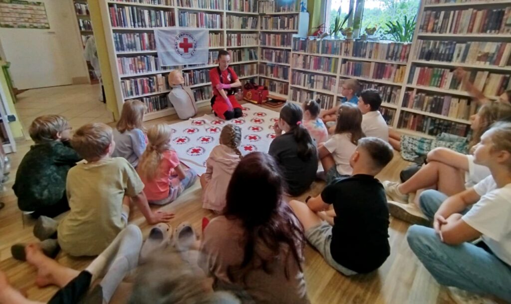 na zdjęciu widać dzieci w sali bibliotecznej uczestniczących w warsztatach pierwszej pomocy. Dzieci siedzą na podłodze i słuchają pani prowadżcej wykład