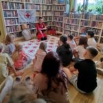 na zdjęciu widać dzieci w sali bibliotecznej uczestniczących w warsztatach pierwszej pomocy. Dzieci siedzą na podłodze i słuchają pani prowadżcej wykład