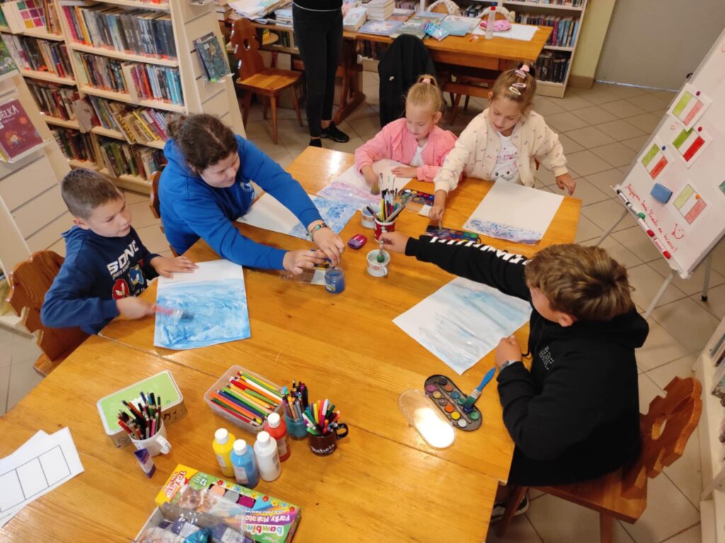 Trzy dziewczynkii dwóch chłopców siedzi przy stole i wykonują pracę plastyczną. na stole kredki, farby, flamastry. Wokół regały z książkami.