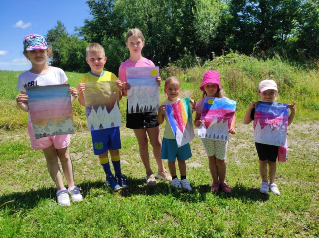 Na zdjęciu na zielonej trawie stoja dzieci pięć dziewczynek i jeden chłopiec. W reku trzymaja kolorowe prace plastyczne wykonane przez siebie.