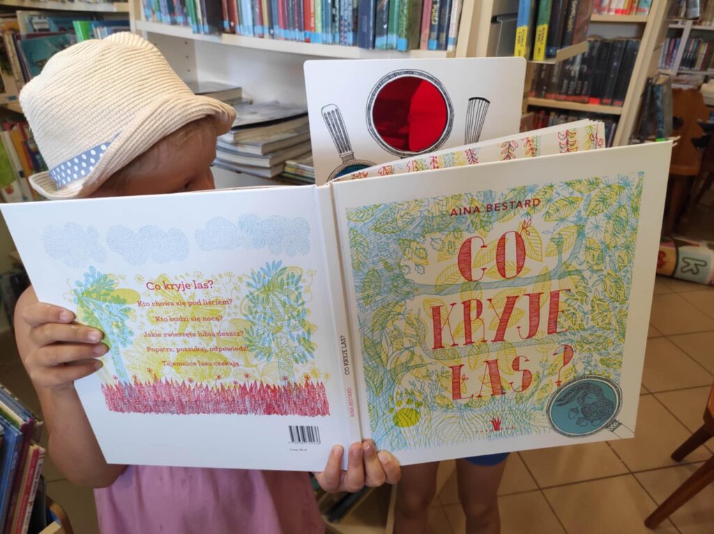 Na zdjęciu dziewczynka,której nie widac twarzy. Trzyma w otwartą książkę, którą przysłania swoja twarz.