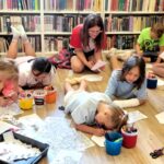 na zdjęciu widać dzieci w sali bibliotecznej rysujące siedząc na podłodze i wykonujące prace plastyczne