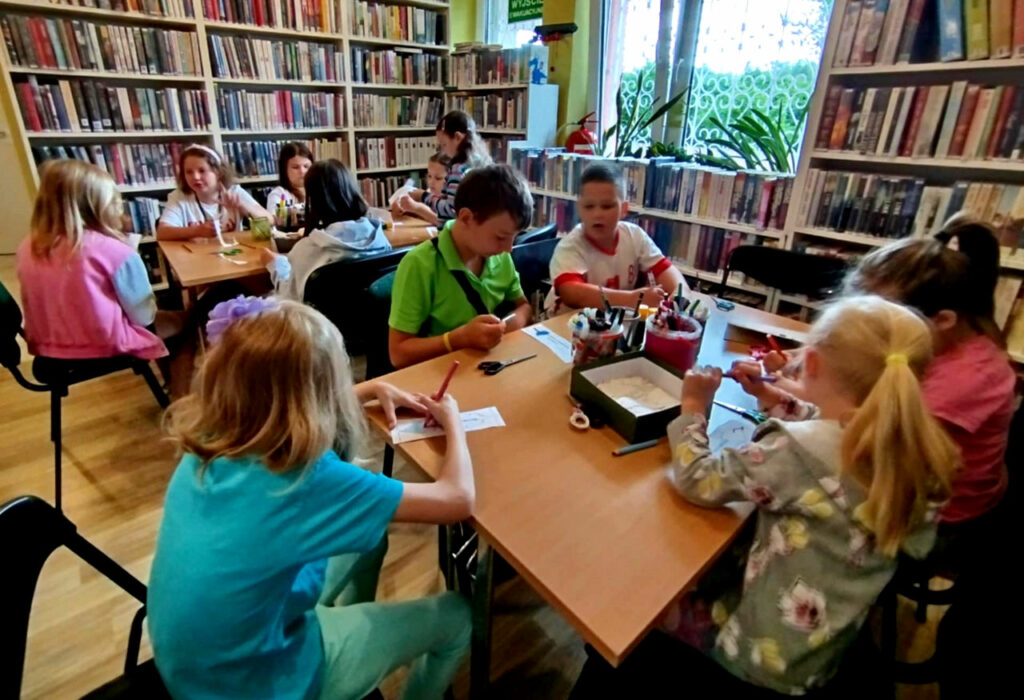 na zdjęciu widać dzieci w sali bibliotecznej siedzące przy stołach i wykonujące prace plastyczne. Na stole kolorowe kredki