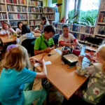 na zdjęciu widać dzieci w sali bibliotecznej siedzące przy stołach i wykonujące prace plastyczne. Na stole kolorowe kredki