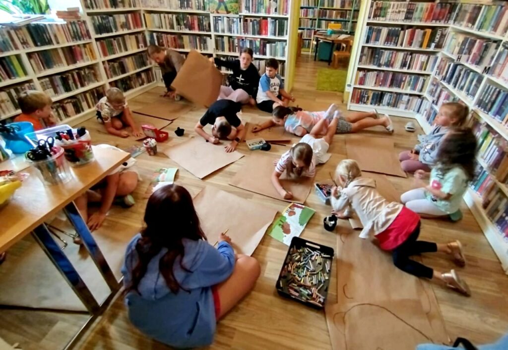 Na zdjęciu dzieci w sali bibliotecznej , na podłodze rozłożone szare papiery. Dzieci wykonują prace plastyczną. Wokół regały z książkami.