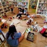 Na zdjęciu dzieci w sali bibliotecznej , na podłodze rozłożone szare papiery. Dzieci wykonują prace plastyczną. Wokół regały z książkami.