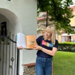 Na zdjęciu bibliotekarka trzyma w ręku książkę i ją prezentuje. Za nią kapliczka.