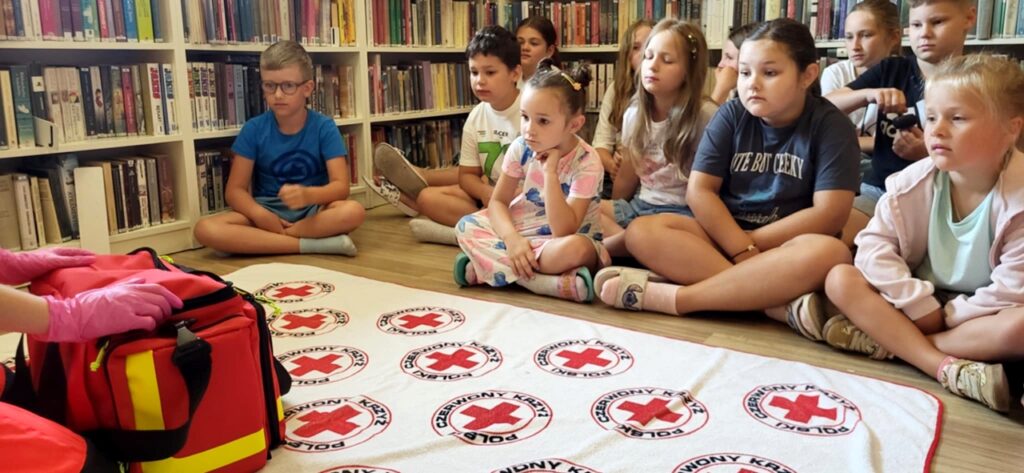na zdjęciu widać dzieci w sali bibliotecznej uczestniczących w warsztatach pierwszej pomocy. Dzieci siedzą na podłodze i słuchaja pani prowadzącej