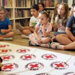 na zdjęciu widać dzieci w sali bibliotecznej uczestniczących w warsztatach pierwszej pomocy. Dzieci siedzą na podłodze i słuchaja pani prowadzącej