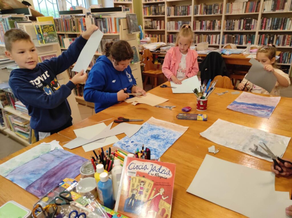 Trzy dziewczynki i chłopiec siedzą i wykonuja prace plastyczną. Na stole kredki, flamastry i nożyczki