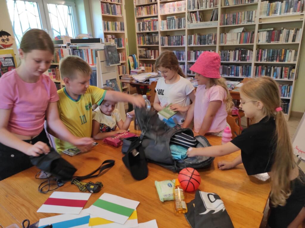 Na zdjęciu szóstka dzieci, która wykonuje zadania zadane przez bibliotekarkę. Na stole plezak, piłka, kolorowe kartki. Wokół regały z książkami.