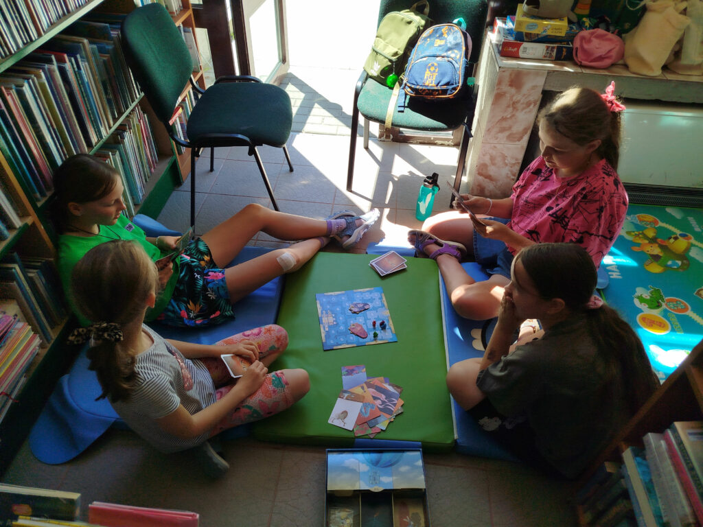 Na zdjęciu widać czter dziewczynki, które siedza na podłodze i grają w gry planszowe. Za nimi regał z ksiązkami, dwa krzesła na których są plecaki