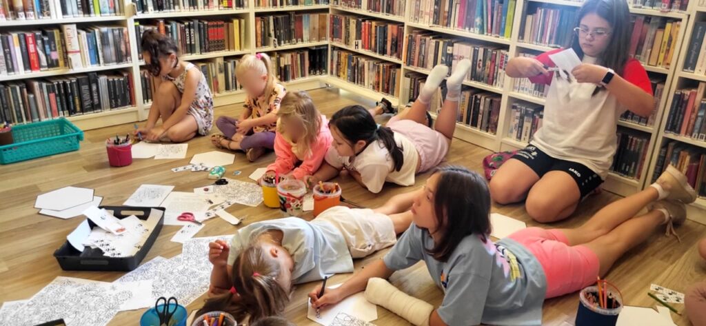 na zdjęciu widać dzieci w sali bibliotecznej rysujące siedząc na podłodze. Malują, wycinają i wykonują prace plastyczne.