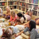 na zdjęciu widać dzieci w sali bibliotecznej rysujące siedząc na podłodze. Malują, wycinają i wykonują prace plastyczne.