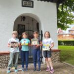 Cztery dziewczynki stoją przed kapliczką. W ręku trzymają wykonane przez siebie prace. Kolorowe obracki z kapliczką w Tyliczu.