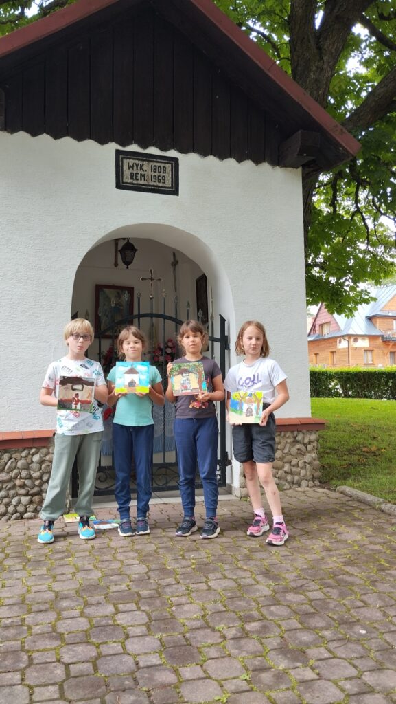 Cztery dziewczynki stoją przed kapliczką. W ręku trzymają wykonane przez siebie prace. Kolorowe obracki z kapliczką w Tyliczu.